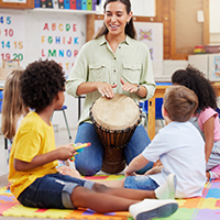 Adult female of color playing a drum for a diverse group of children in a classroom.