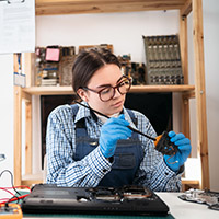 Person with overalls and glasses cleaning an electrical component in a workshop.