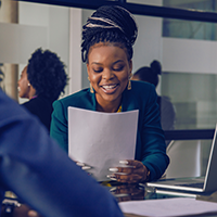 Decorative image of a woman sitting behind a desk, smiling as she looks at a sheet of paper.