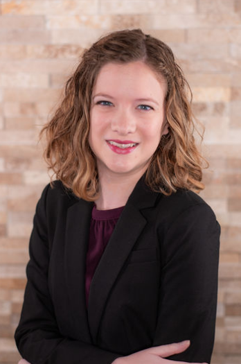 Woman with dark curly hair smiling at camera. She is wearing a black blazer with a burgundy top.