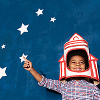 Male BIPOC child, wearing a paper mache helmet made to look like a rocketship, pointing to white stars on a blue background.