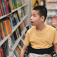 Asian child with disability in wheelchair looking at library books. 